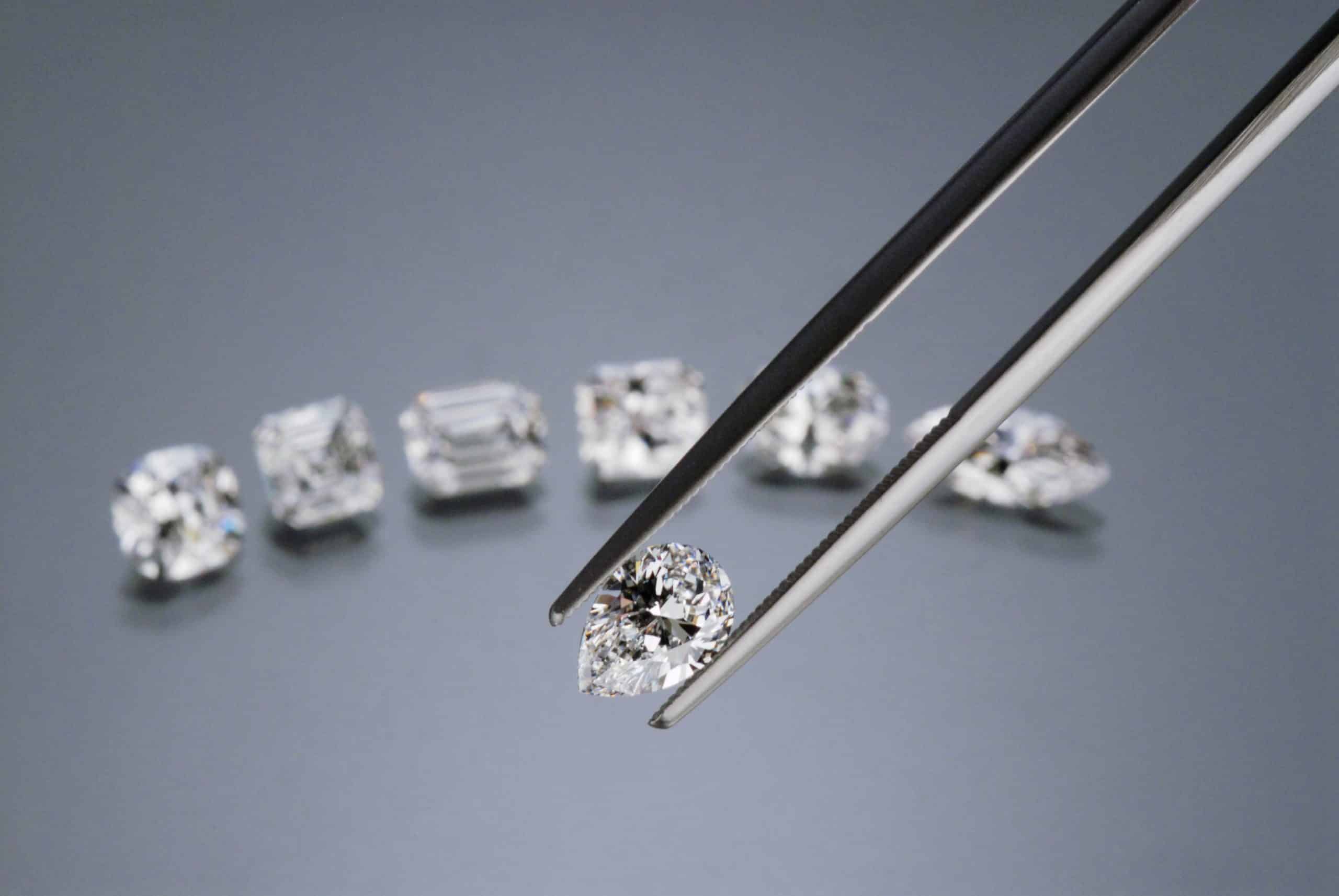 A collection of loose diamonds in various shapes, including round, emerald, and pear cuts, being handled with professional jeweler tweezers